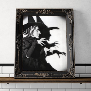 May include: Black and white photo of a woman in a witch costume with a pointed hat and a long dress. The woman is making a gesture with her hands, and her shadow is cast on the wall behind her.