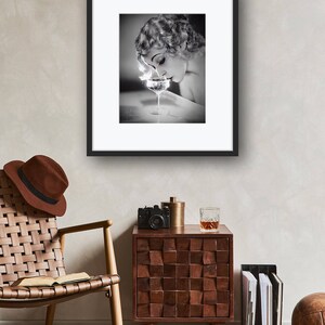 Op de afbeelding: Een zwart-witfoto van een vrouw in een vintage jurk, die aan een glas champagne ruikt. De foto is ingelijst in een zwart frame en hangt aan een muur.