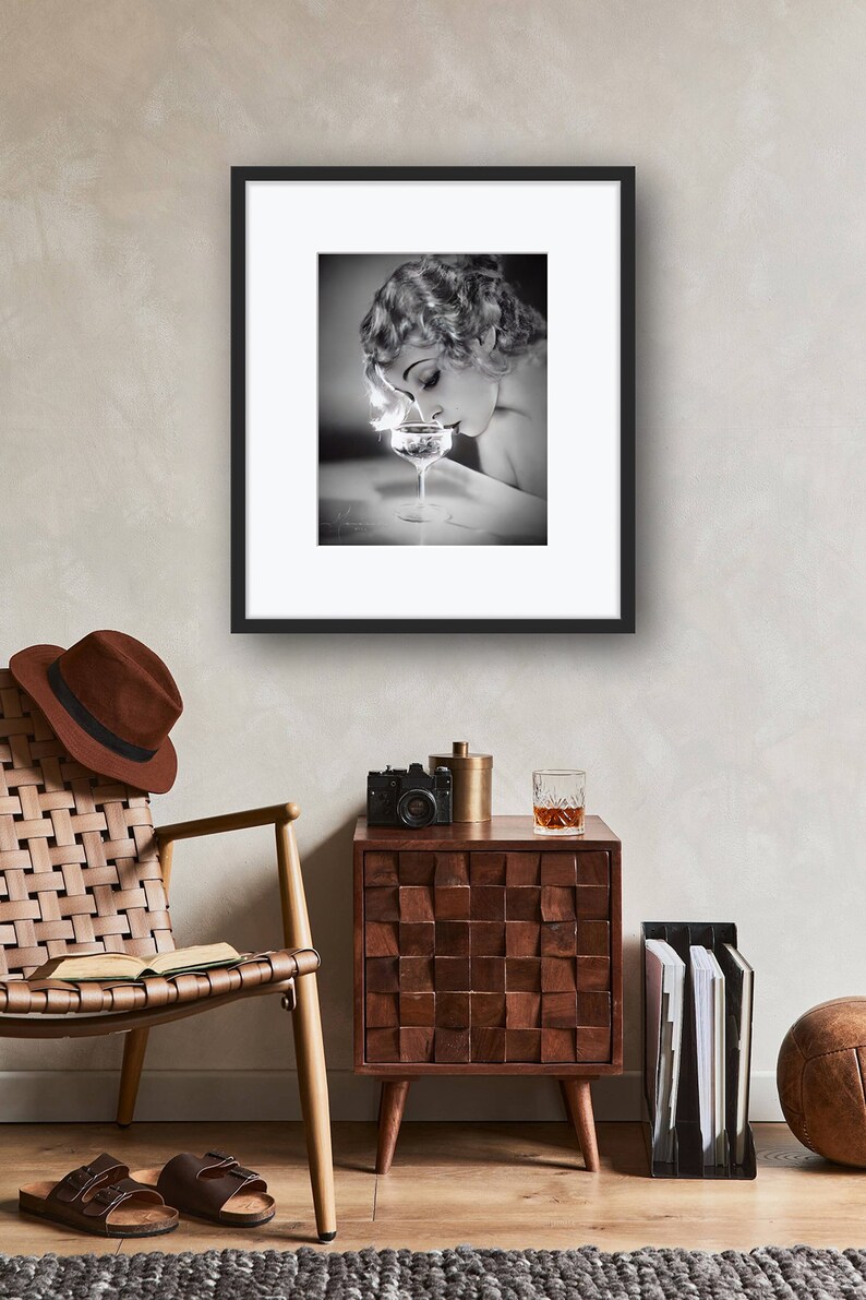 Op de afbeelding: Een zwart-witfoto van een vrouw in een vintage jurk, die aan een glas champagne ruikt. De foto is ingelijst in een zwart frame en hangt aan een muur.