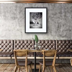 Op de afbeelding: Een zwart-witfoto van een vrouw in een vintage setting, die naar beneden kijkt naar een glas vloeistof. De foto is ingelijst en hangt aan een muur in een restaurant.