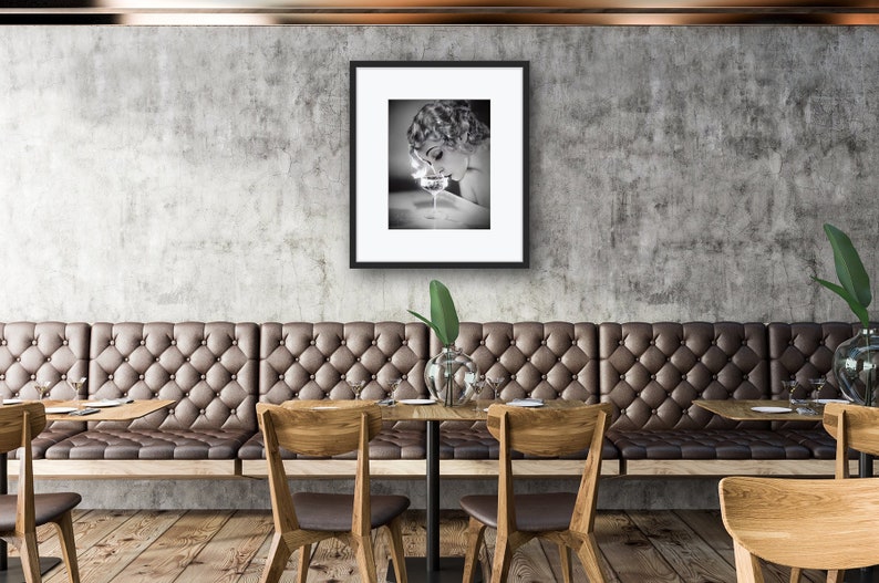 Op de afbeelding: Een zwart-witfoto van een vrouw in een vintage setting, die naar beneden kijkt naar een glas vloeistof. De foto is ingelijst en hangt aan een muur in een restaurant.