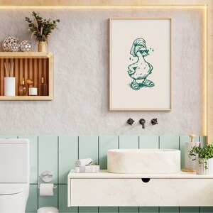 Pu&ograve; includere: Una stampa incorniciata con un'anatra dei cartoni animati con occhiali da sole, un asciugamano in testa e sandali. L'anatra &egrave; disegnata con inchiostro verde su sfondo bianco sporco. La stampa &egrave; esposta in un bagno con cornice in legno chiaro.