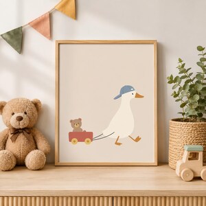 May include: A framed wall art print featuring a white duck wearing a blue baseball cap, pulling a red wagon that contains a small brown teddy bear. The illustration is set against a solid light beige background within a simple wooden frame.