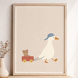 May include: A framed wall art print featuring a white duck wearing a blue baseball cap, pulling a small red wagon that contains a brown teddy bear. The illustration is set against a solid light beige background within a light wood frame.