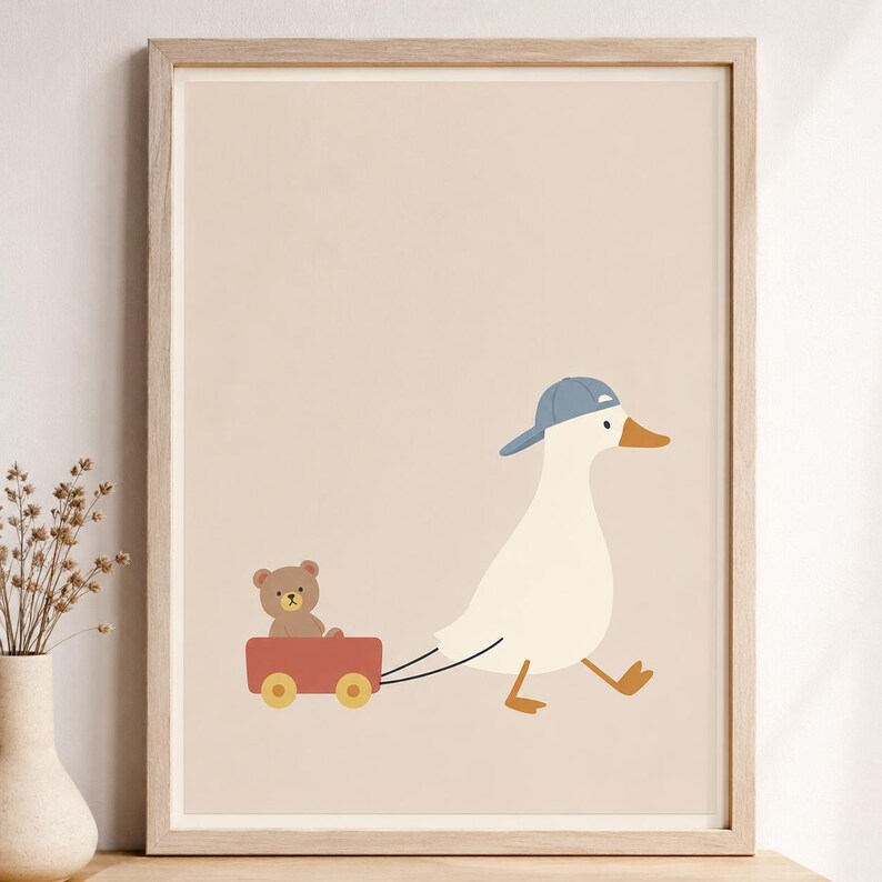 May include: A framed wall art print featuring a white duck wearing a blue baseball cap, pulling a small red wagon that contains a brown teddy bear. The illustration is set against a solid light beige background within a light wood frame.