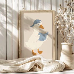 May include: A framed print featuring a cartoon goose wearing a blue baseball cap and reading a blue book. The goose is white with an orange beak and feet. The background is a light beige color.