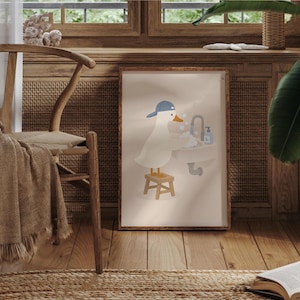 May include: A framed wall art print featuring a white cartoon duck wearing a blue baseball cap, standing on a small wooden stool while washing its wings in a bathroom sink with a faucet and soap dispenser, set against a light beige background.