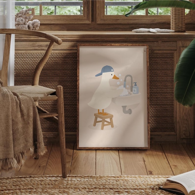 May include: A framed wall art print featuring a white cartoon duck wearing a blue baseball cap, standing on a small wooden stool while washing its wings in a bathroom sink with a faucet and soap dispenser, set against a light beige background.