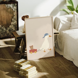 Pode incluir: Impress&atilde;o emoldurada com uma ilustra&ccedil;&atilde;o de um pato de desenho animado com bon&eacute; azul puxando uma carro&ccedil;a vermelha com um ursinho de pel&uacute;cia. A obra tem um fundo bege claro e uma moldura de madeira, ideal para um quarto infantil.
