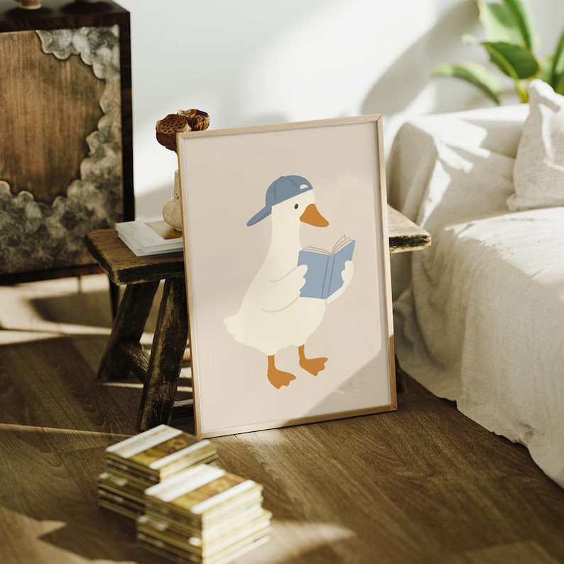 May include: A framed art print featuring a cartoon goose wearing a blue baseball cap and reading a blue book. The goose is white with orange feet and a beak. The background is a light beige color.