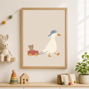 May include: A framed wall art print featuring a white duck wearing a blue baseball cap, pulling a red wagon that contains a small brown teddy bear. The illustration is set against a solid light beige background and enclosed in a light wood frame.