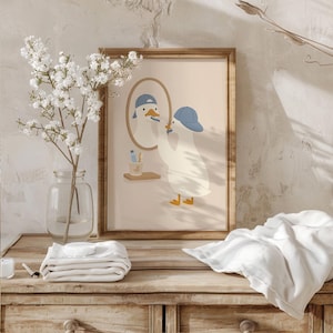 May include: A framed wall art print featuring a cartoon duck wearing a blue baseball cap, standing in front of an oval mirror while brushing its beak. The illustration includes a small shelf with a toothbrush holder and a toothbrush in a soft beige tone.