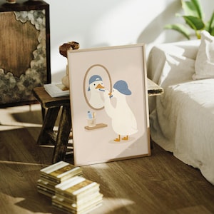 May include: A framed wall art print featuring a white duck wearing a blue baseball cap, standing in front of an oval mirror while brushing its beak with a blue toothbrush. A small shelf below the mirror holds a blue tube and a wooden toothbrush.