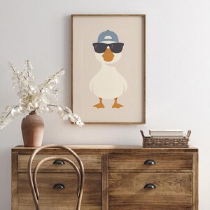 May include: A framed art print featuring a cartoon goose wearing sunglasses and a blue baseball cap. The goose has an orange beak and feet, set against a neutral background. The artwork is in a wooden frame.