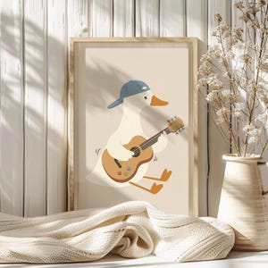 May include: A framed art print featuring a cartoon goose wearing a blue baseball cap and playing a brown acoustic guitar. The goose is depicted with a white body, orange beak and feet, set against a beige background. The frame is a light wood color.