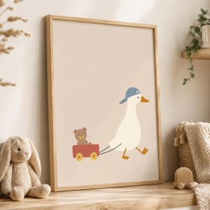 May include: A framed wall art print featuring a white duck wearing a blue baseball cap, pulling a small red wagon that contains a brown teddy bear. The illustration is set against a solid light beige background, presented in a light wood frame.