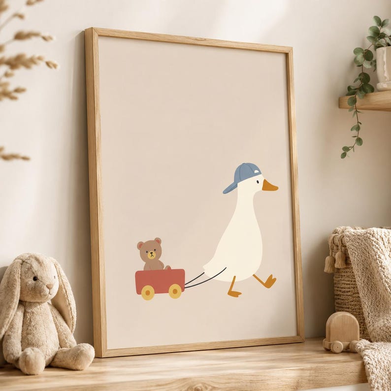 May include: A framed wall art print featuring a white duck wearing a blue baseball cap, pulling a small red wagon that contains a brown teddy bear. The illustration is set against a solid light beige background, presented in a light wood frame.