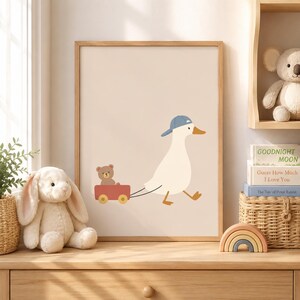May include: A framed wall art print featuring a white duck wearing a blue baseball cap, pulling a small red wagon that carries a brown teddy bear. The illustration is set against a solid light beige background, presented in a wooden frame.