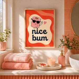Op de afbeelding: Een rechthoekige wandprint in een lichthouten lijst met een cartoon wc-rol die een zwarte zonnebril draagt en een vredesteken maakt. De tekst "nice bum" staat in donkerblauw gedrukt tegen een golvende rode en cr&egrave;mekleurige achtergrond.
