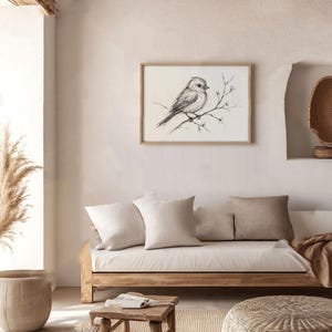 Op de afbeelding: Een zwart-witte schets van een vogel die op een tak zit met kleine witte bloemen. De schets is ingelijst en hangt aan een witte muur boven een lichtbruine bank met witte kussens.