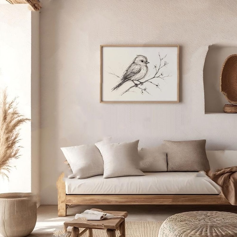 Op de afbeelding: Een zwart-witte schets van een vogel die op een tak zit met kleine witte bloemen. De schets is ingelijst en hangt aan een witte muur boven een lichtbruine bank met witte kussens.