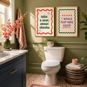 Op de afbeelding: Twee rechthoekige wandposters in licht houten lijsten. De linker poster heeft een golvende rode rand, groene tekst met "take a seat, sweet cheeks" en twee kleine paarse sterren. De rechter poster heeft een golvende groene rand, vijf rode sterren en paarse tekst met "I WOULD POOP HERE AGAIN" met een roze onderstreping.