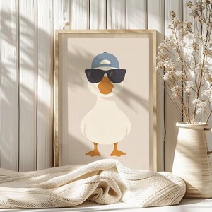 May include: A framed print featuring a cartoon duck wearing sunglasses and a blue baseball cap. The duck has a white body, an orange beak and feet, and is set against a neutral background. The artwork is displayed on a white wooden surface.