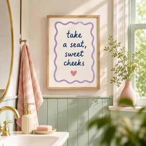 May include: A rectangular wall art print in a light wood frame featuring a wavy purple border on a white background. The text reads "take a seat, sweet cheeks" in dark blue script, with a small pink heart centered below the text.