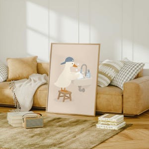 May include: A framed wall art print featuring a white duck wearing a blue baseball cap, standing on a small wooden stool while washing its wings in a sink with soap bubbles, a faucet, and a blue soap dispenser against a light beige background.