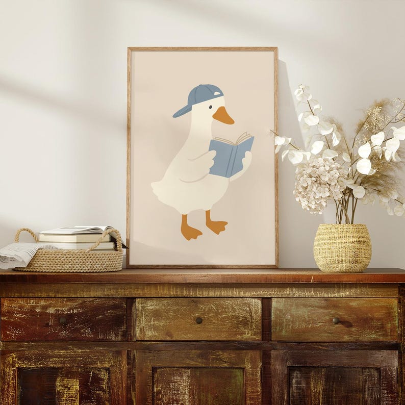 May include: A framed art print featuring a cartoon goose wearing a blue baseball cap and reading a blue book. The goose is white with orange feet and a beak. The background is a light beige color.