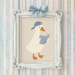 May include: A framed print featuring a cartoon goose wearing a blue baseball cap and reading a blue book. The goose is white with orange feet and a beak. The frame is white with ornate detailing, and a blue bow is attached at the top.