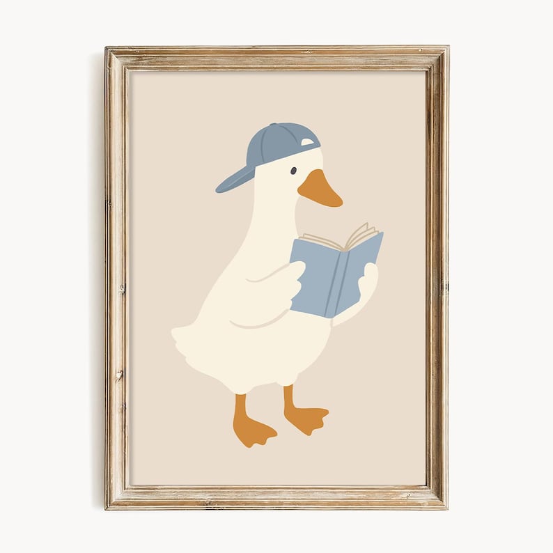 May include: A framed print featuring a cartoon goose wearing a blue baseball cap and reading a blue book. The goose is white with orange feet and a beak, set against a beige background. The frame is a light brown color.