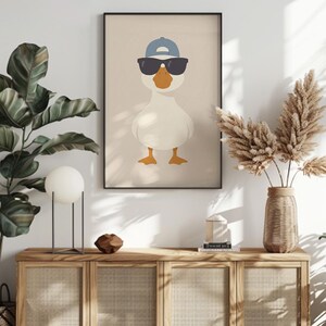 May include: Framed art print featuring a cartoon goose wearing sunglasses and a blue baseball cap. The goose is white with orange feet and beak, set against a neutral background. The artwork is in a black frame.