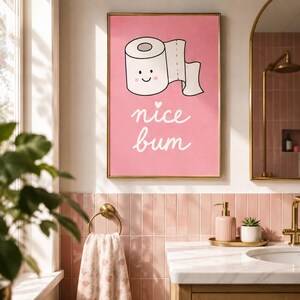 May include: A rectangular wall art print with a pink background featuring a white cartoon toilet paper roll with a smiling face and the white cursive text "nice bum" with a small heart above the word nice, framed in a thin gold metal border.