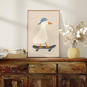 May include: A framed print featuring a cartoon illustration of a white duck wearing a blue baseball cap and riding a black skateboard. The artwork has a beige background and is displayed on a wooden cabinet.
