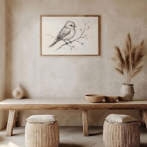 Op de afbeelding: Een zwart-witte inkttekening van een vogel die op een tak zit met kleine bloemen. De tekening is ingelijst en hangt aan een lichtbeige muur. De muur is versierd met een vaas met gedroogd pampasgras en een houten tafel met twee geweven manden.