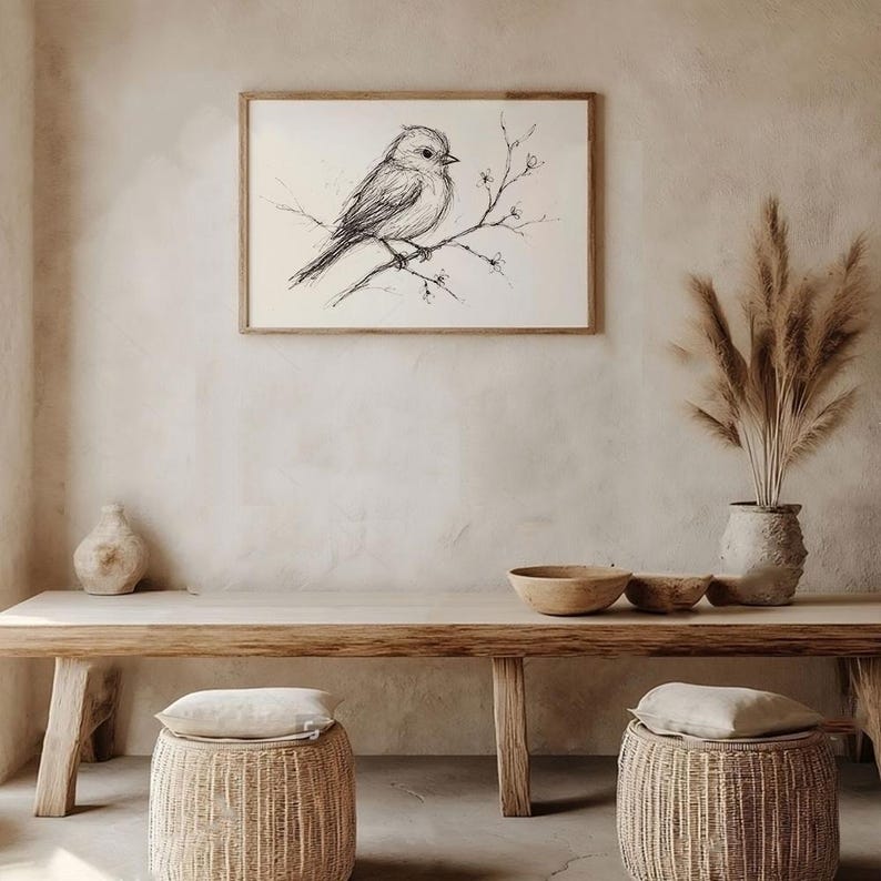 Op de afbeelding: Een zwart-witte inkttekening van een vogel die op een tak zit met kleine bloemen. De tekening is ingelijst en hangt aan een lichtbeige muur. De muur is versierd met een vaas met gedroogd pampasgras en een houten tafel met twee geweven manden.