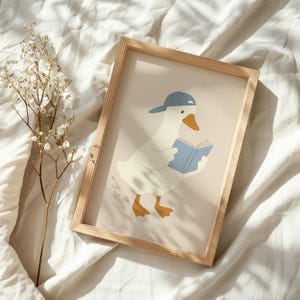 May include: A framed print featuring a cartoon goose wearing a blue baseball cap and reading a blue book. The goose has white feathers, an orange beak and feet. The frame is made of light-colored wood. A sprig of white flowers is to the left.