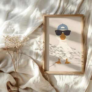 May include: A framed print featuring a cartoon duck wearing sunglasses and a blue baseball cap. The duck has an orange beak and yellow feet. The artwork is set against a neutral background and framed in a light wood.