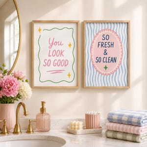 May include: Two rectangular wall art prints in light wood frames. The left print has a white background with a green wavy border, yellow stars, and the pink text "You LOOK SO GOOD". The right print features blue wavy lines, a pink oval, and "SO FRESH & SO CLEAN" in dark blue text.