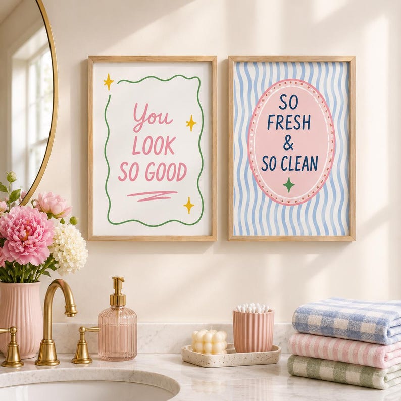 May include: Two rectangular wall art prints in light wood frames. The left print has a white background with a green wavy border, yellow stars, and the pink text "You LOOK SO GOOD". The right print features blue wavy lines, a pink oval, and "SO FRESH & SO CLEAN" in dark blue text.