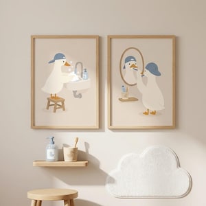 May include: Two framed wall art prints featuring a white duck wearing a blue baseball cap. The left print shows the duck washing hands at a sink on a stool. The right print shows the duck brushing teeth in front of a mirror. Below are a soap dispenser, a toothbrush holder, a wooden stool, and a cloud-shaped rug.