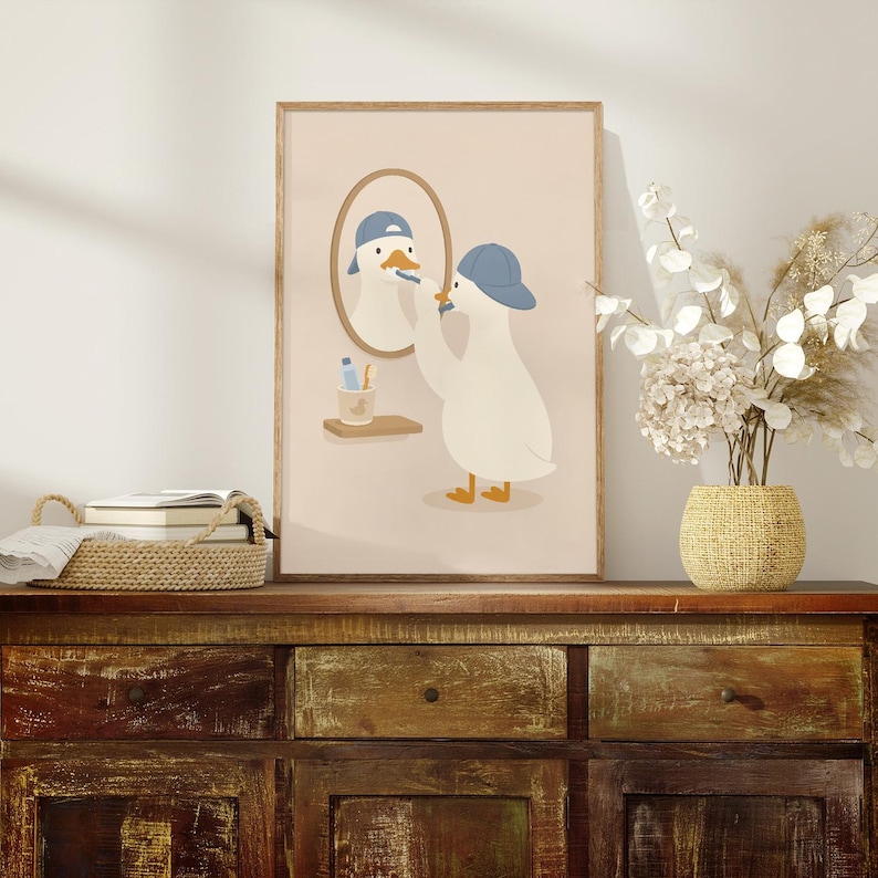 May include: A framed wall art print featuring a white duck wearing a blue baseball cap, brushing its teeth in front of an oval mirror. The duck stands on a light beige background, with a small shelf holding a toothbrush cup visible below the mirror.