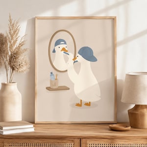 May include: A framed wall art print featuring a white duck wearing a blue baseball cap, standing before an oval mirror while brushing its beak with a toothbrush. A small shelf holds a blue bottle and a toothbrush in a cup with a duck silhouette.