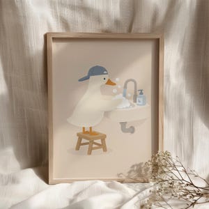 May include: A framed wall art print featuring a white duck wearing a blue baseball cap, standing on a small wooden stool while washing its wings in a bathroom sink with soap bubbles, a faucet, and a blue soap dispenser on a beige background.