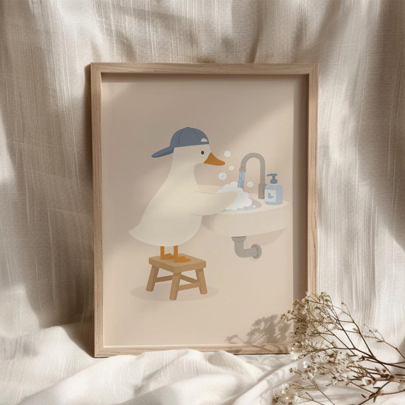 May include: A framed wall art print featuring a white duck wearing a blue baseball cap, standing on a small wooden stool while washing its wings in a bathroom sink with soap bubbles, a faucet, and a blue soap dispenser on a beige background.
