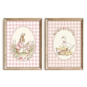 May include: Two framed prints with watercolour illustrations. One shows a rabbit and a smaller rabbit in pink jackets, surrounded by flowers. The other depicts a white goose with a pink bow and ducklings in a basket, with floral accents.