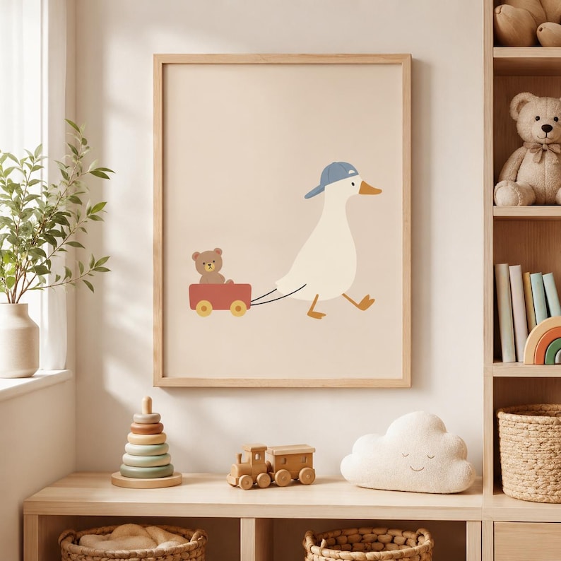 May include: A framed wall art print featuring a white duck wearing a blue baseball cap, pulling a red wagon with a small brown teddy bear inside. The illustration is set against a solid light beige background, presented in a light wood frame.