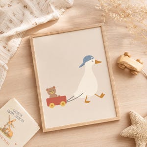 May include: A framed wall art print featuring a white duck wearing a blue baseball cap, pulling a red wagon that carries a small brown teddy bear. The illustration is set against a solid light beige background, presented in a light wood frame.