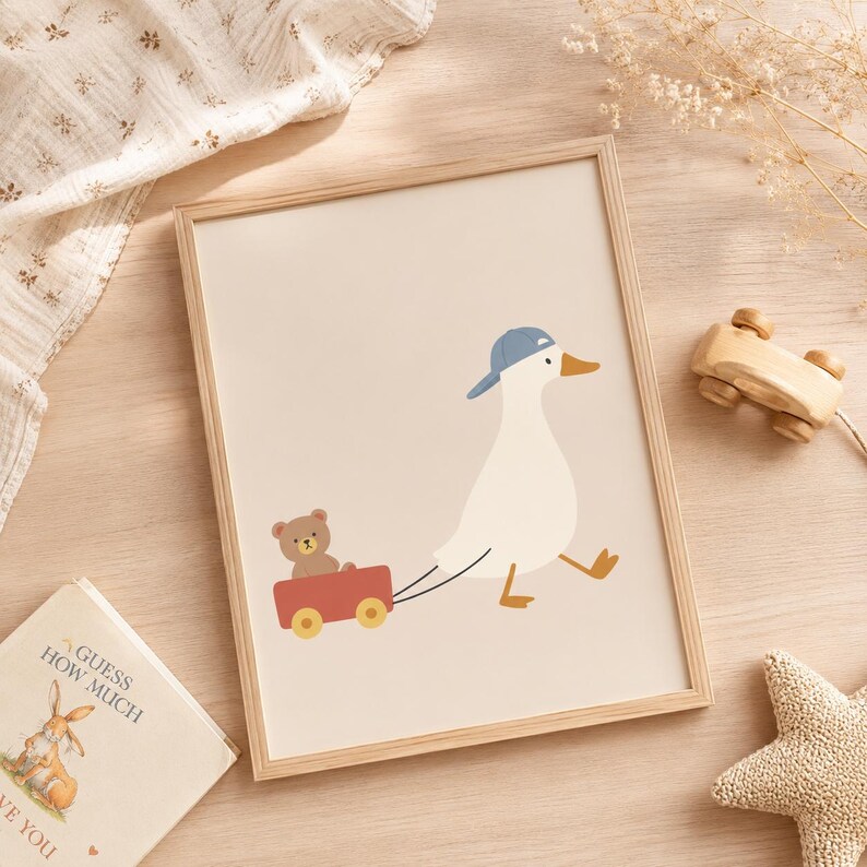 May include: A framed wall art print featuring a white duck wearing a blue baseball cap, pulling a red wagon that carries a small brown teddy bear. The illustration is set against a solid light beige background, presented in a light wood frame.